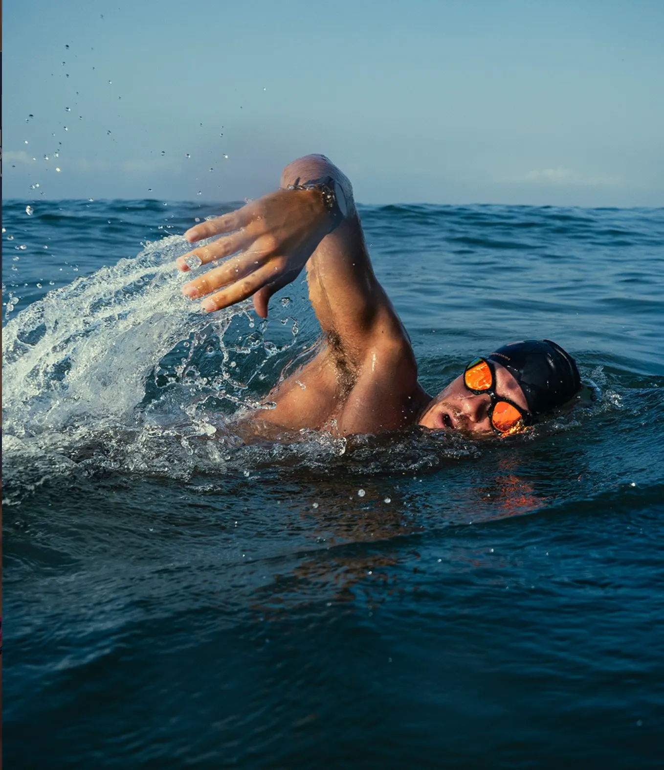 Open-water swimmer mid-stroke in the sea, wearing Buddyswim goggles with orange mirrored lenses