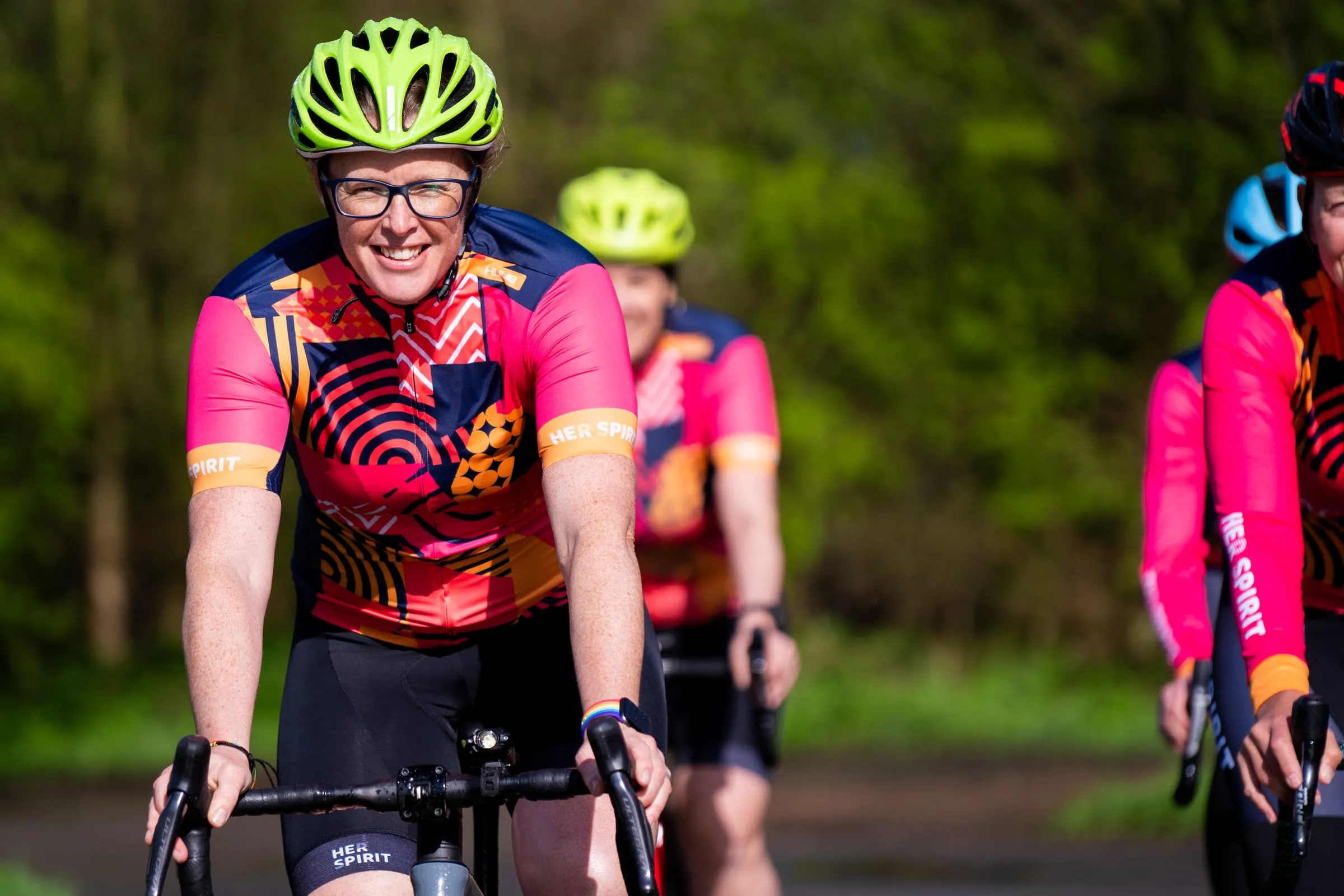 A smiling cyclist in a HUUB Her Spirit jersey leading a group ride along a leafy lane