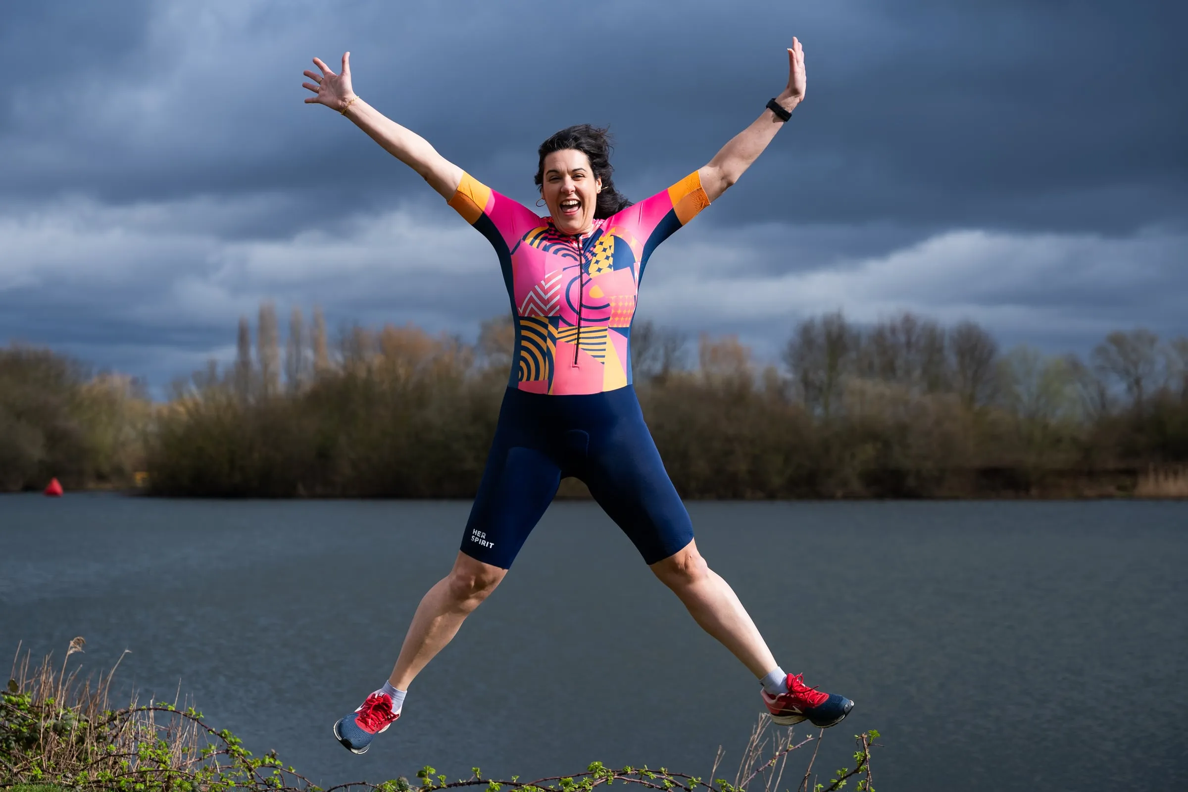 A woman leaping in a star jump beside a lake in a HUUB Her Spirit trisuit under a stormy sky